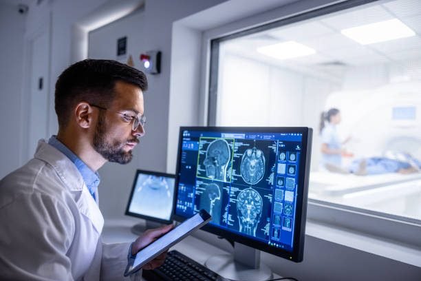 Doctor examining X-ray images on display in control room while in background nurse preparing the patient for new MRI scanning test.